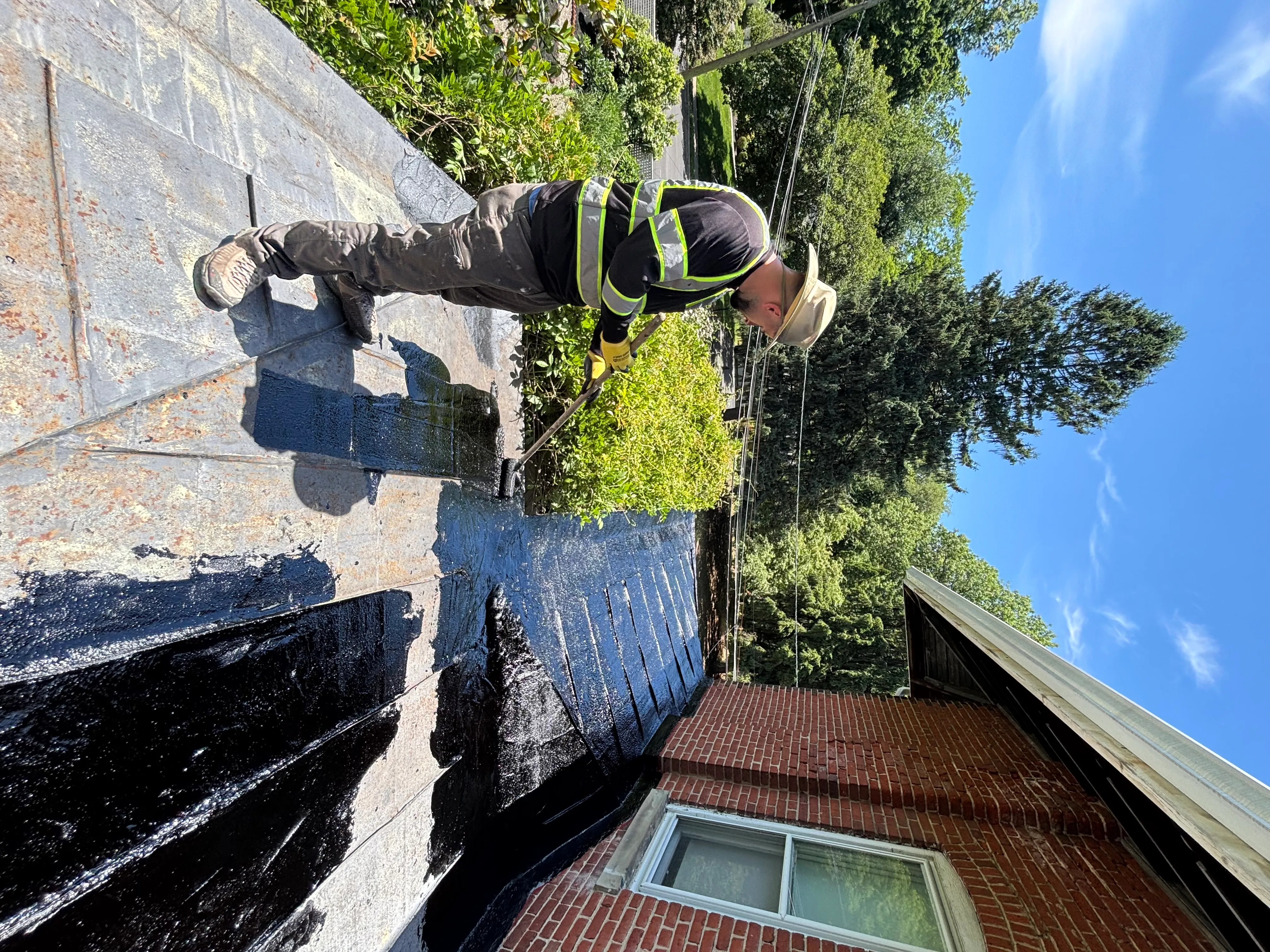 Roofer in a hi-vis vest on a ladder brushing black roof coating onto the parapet under a clear blue sky.