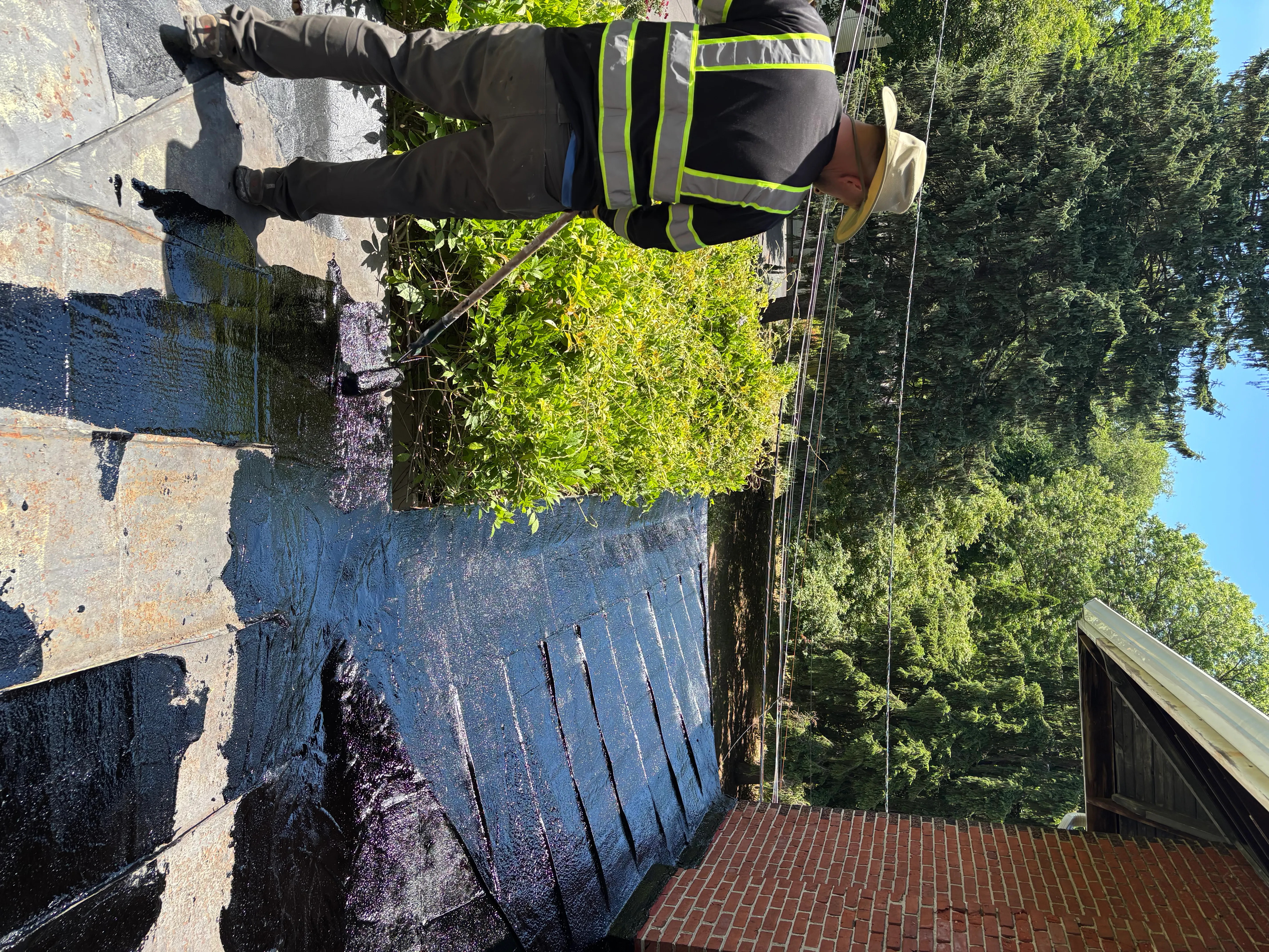 Roofer applying black coating to the parapet, half the surface coated glossy black and half still bare.