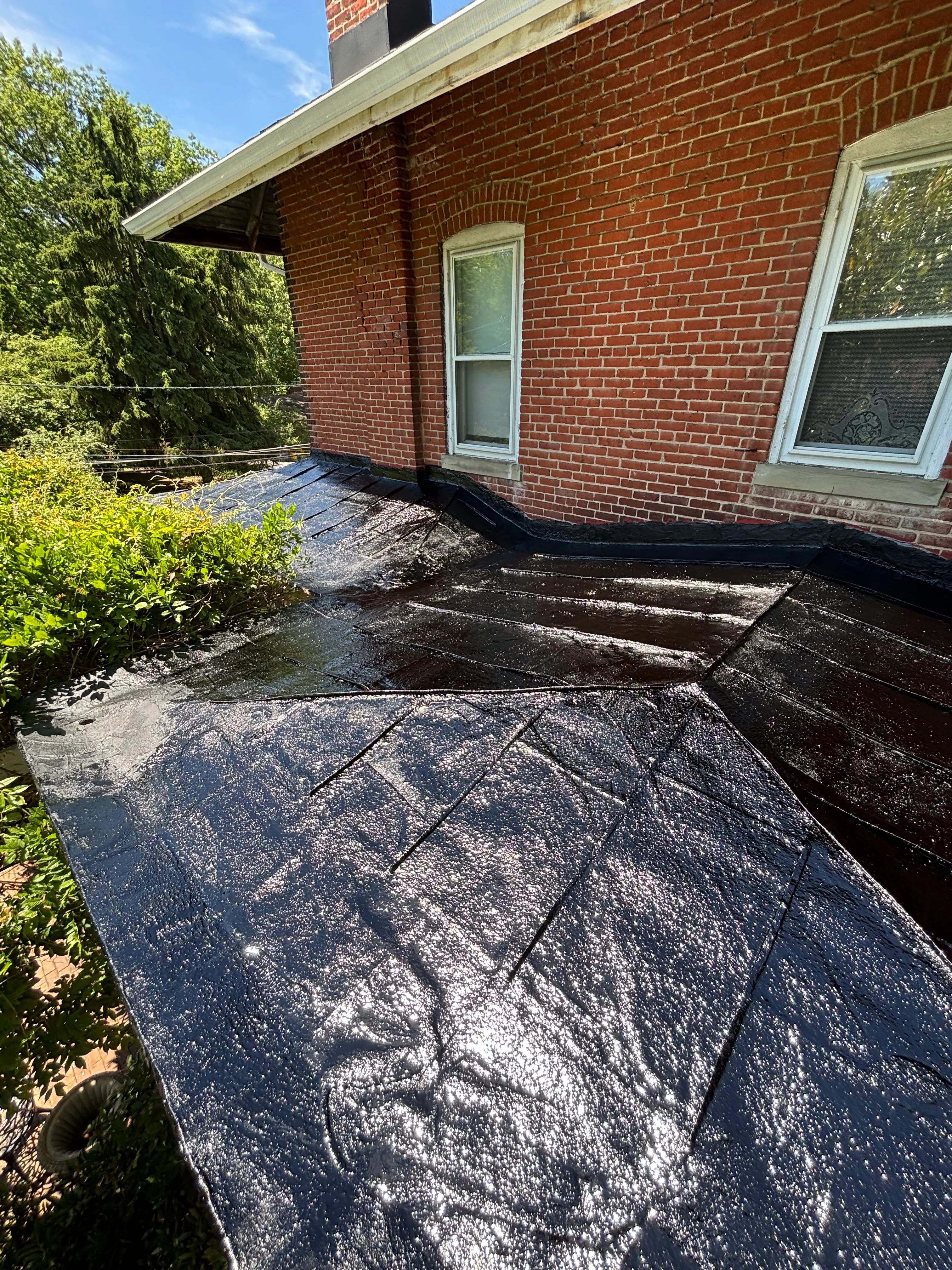 Finished black-coated flat roof meeting the brick house wall and windows, the new coating glossy and even.