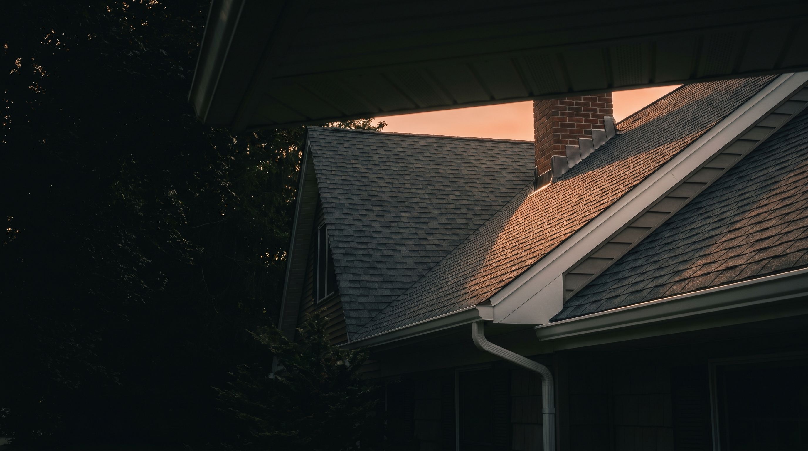 A completed asphalt-shingle roof on a North Jersey colonial at golden hour, with new flashing at the brick chimney and white gutters along the eave.